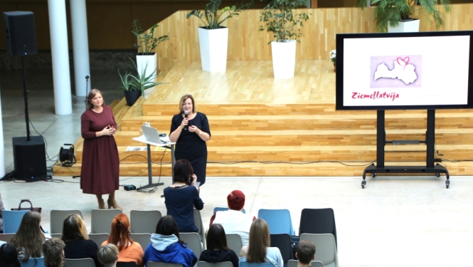 foto ar sievietem kas runā auditorijas priekšā, blakus ekrāns ar uzrakstu ziemeļlatvija