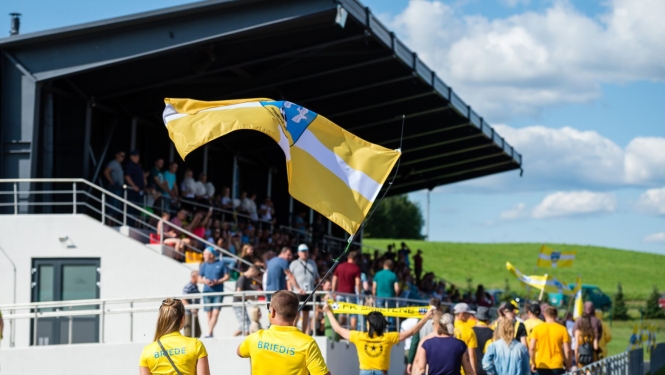 foto ar tepera stadionu , novada karogs fonā un cilvēki 