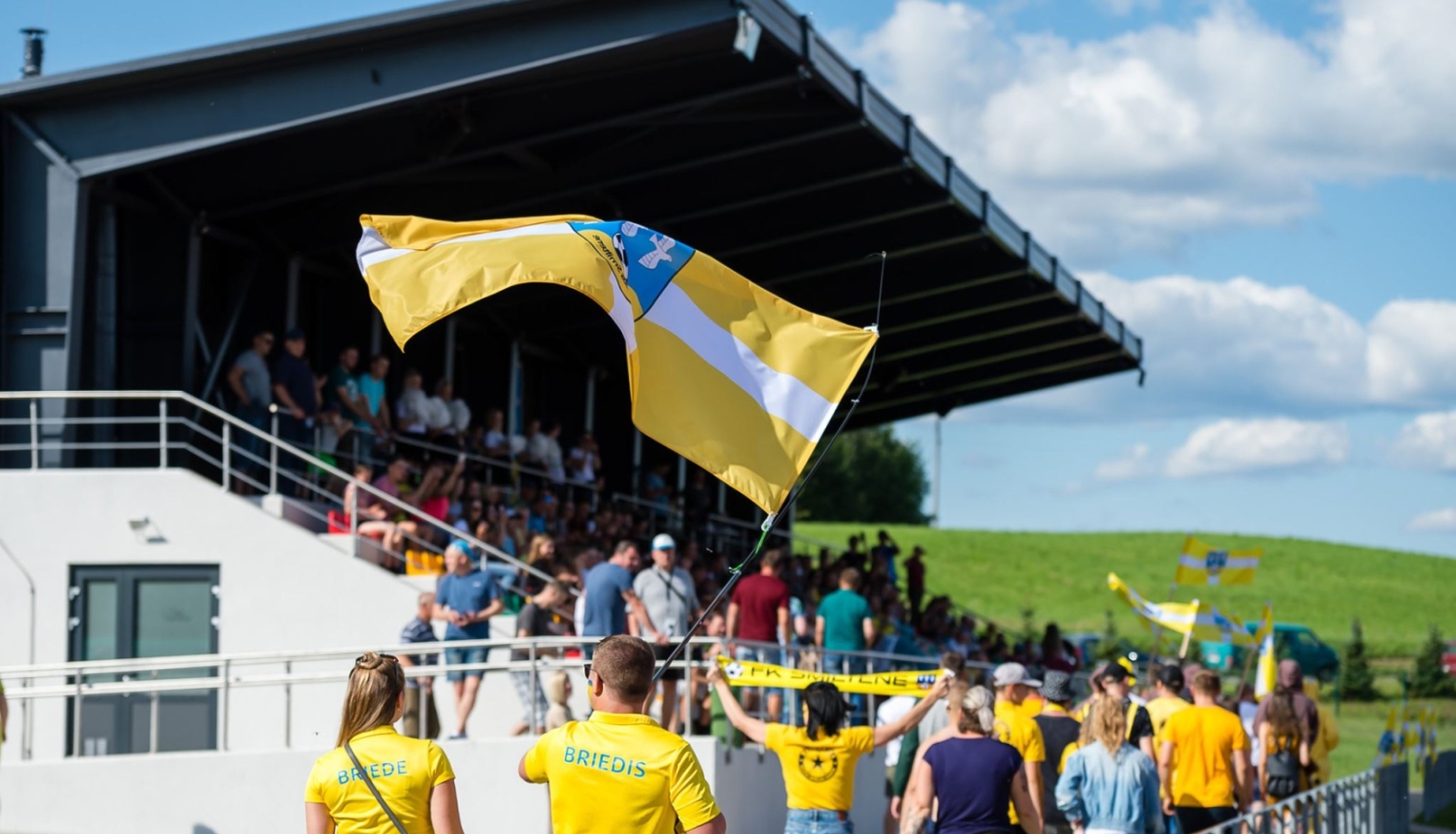 foto ar tepera stadionu , novada karogs fonā un cilvēki 