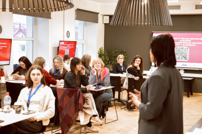 Sieviete stāv grupas priekšā, pārliecinoši prezentējot auditorijai savas idejas un informāciju.
