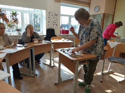 Foto ar skolotajiem pie galda, viens demonstrē kā pērļo rotas