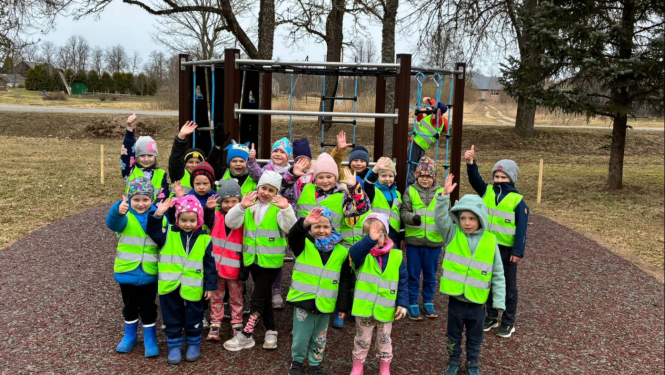 bērni ar dzeltenām vestēm stāv un māj ar roku pie rotaļu objekta 