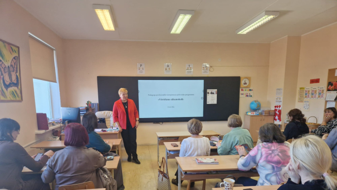 Sieviete klasē vada prezentāciju pie interaktīvās tāfeles, kamēr pieaugušie klausītāji sēž pie soliem.