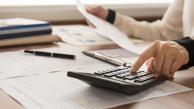 Cilvēks izmanto kalkulatoru un tur dokumentus pie galda, kas noklāts ar papīriem, planšeti un pildspalvu.