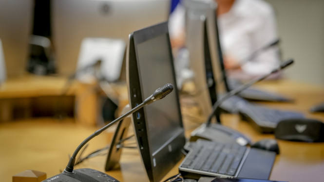 Konferenču mikrofonu, datoru monitoru, tastatūru un cita biroja aprīkojuma tuvplāns uz sanāksmju galda.