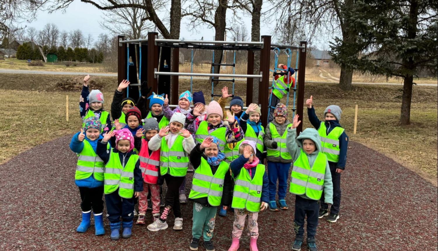 bērni ar dzeltenām vestēm stāv un māj ar roku pie rotaļu objekta 