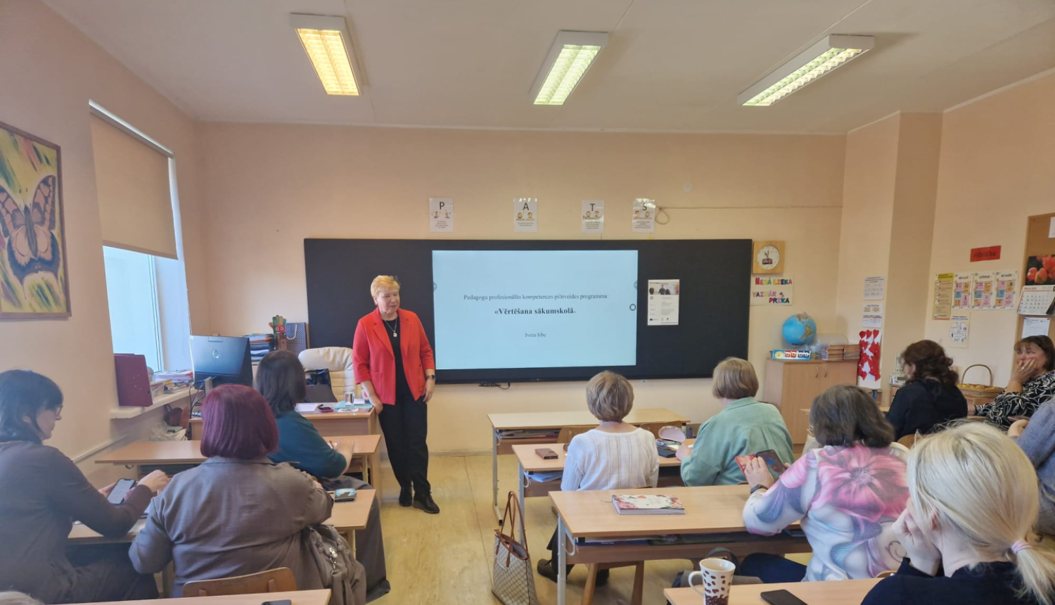 Sieviete klasē vada prezentāciju pie interaktīvās tāfeles, kamēr pieaugušie klausītāji sēž pie soliem.