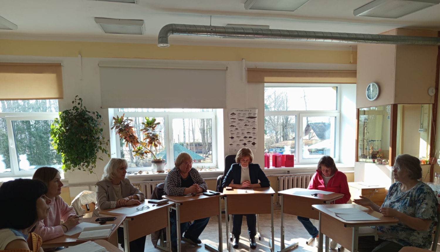 attēlā skolotāji sēž aplī pie galda 